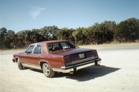 1986 Ford Crown Victoria LTD - A red sedan is parked on a dirt lot with trees in the background.