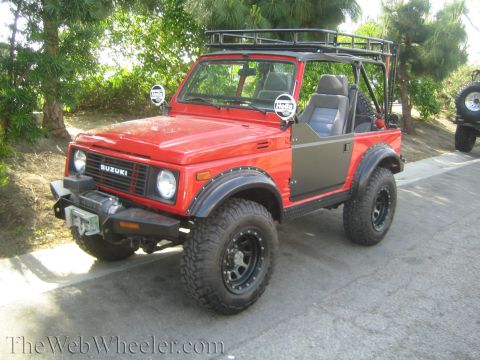 1987 Suzuki Samurai - A red 1987 Suzuki Samurai is parked on a paved surface with trees in the background, and another vehicle is partially visible to the right.