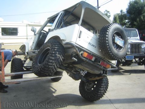 1987 Suzuki Samurai - A modified Suzuki Samurai is being lifted, revealing its suspension and large tires, with a Jeep in the background.