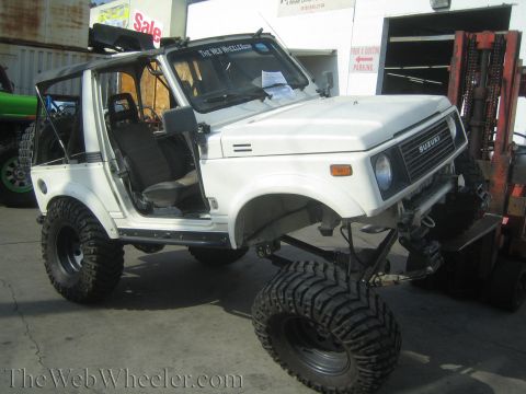 1987 Suzuki Samurai - A white 1987 Suzuki Samurai is shown with a lifted suspension and oversized tires, parked near a forklift.