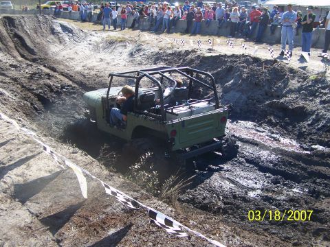 1989 Ford Bronco - A Jeep navigates a muddy obstacle course as spectators watch from the sidelines.