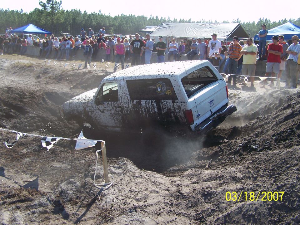 White SUV stuck in a mud pit, covered in mud, with spectators watching