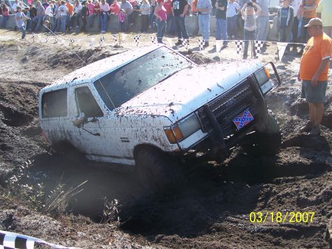 1989 Ford Bronco - A white SUV is stuck in a mud pit during an event, with spectators watching from the sidelines.