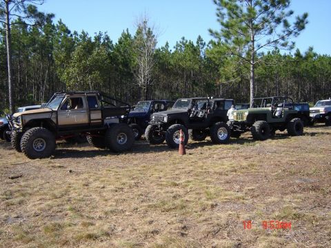 1989 Ford Bronco - Several off-road vehicles are lined up in a grassy field, with a modified pickup truck in the foreground.