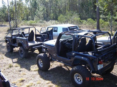1989 Ford Bronco - Modified off-road vehicles are parked in a grassy, wooded area on a sunny day.