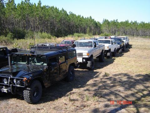 1989 Ford Bronco - A convoy of modified off-road vehicles, including a Humvee and pickup trucks, are lined up outdoors.