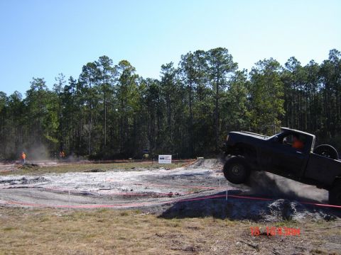 1989 Ford Bronco - Off-road truck catches air over a dirt ramp during an event.