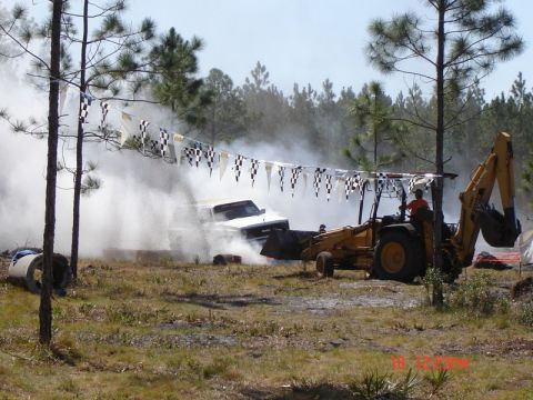 1989 Ford Bronco - A white pickup truck is obscured by smoke during an event, with a backhoe nearby and checkered flags strung overhead.