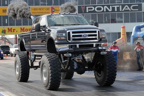 1996 Ford Bronco - A heavily modified black Ford truck with massive tires spews dark smoke during an event.