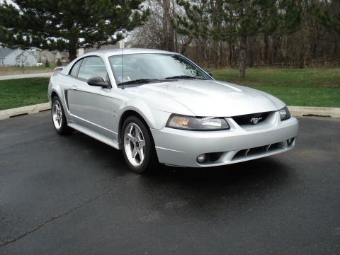 1998 Ford Taurus - This silver Ford Mustang is parked outside on a paved surface with trees and houses in the background.