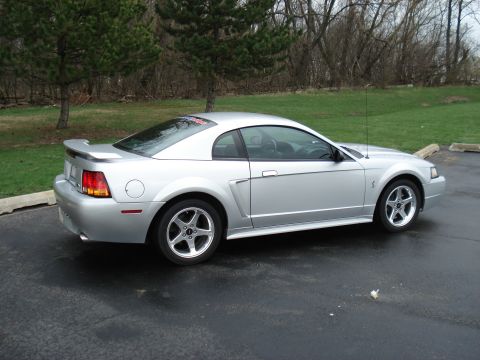 1998 Ford Taurus - A silver coupe with a spoiler and distinctive wheels is parked on a wet asphalt surface.