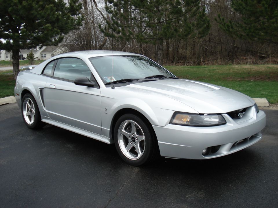 Silver Ford Mustang parked on asphalt, trees and houses in background