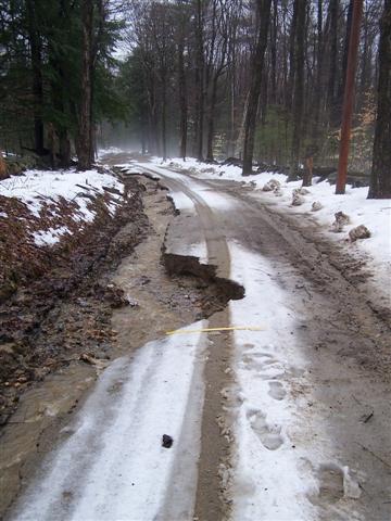 1966 Land Rover Discovery - A dirt road in a forest is partially covered in snow and ice, with a significant section eroded away from the camera eroded.