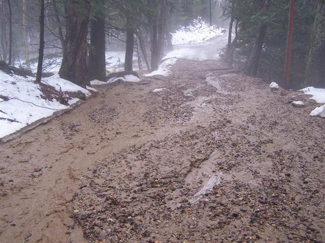 Muddy and rocky forest road with snow on the sides, water flowing down the middle