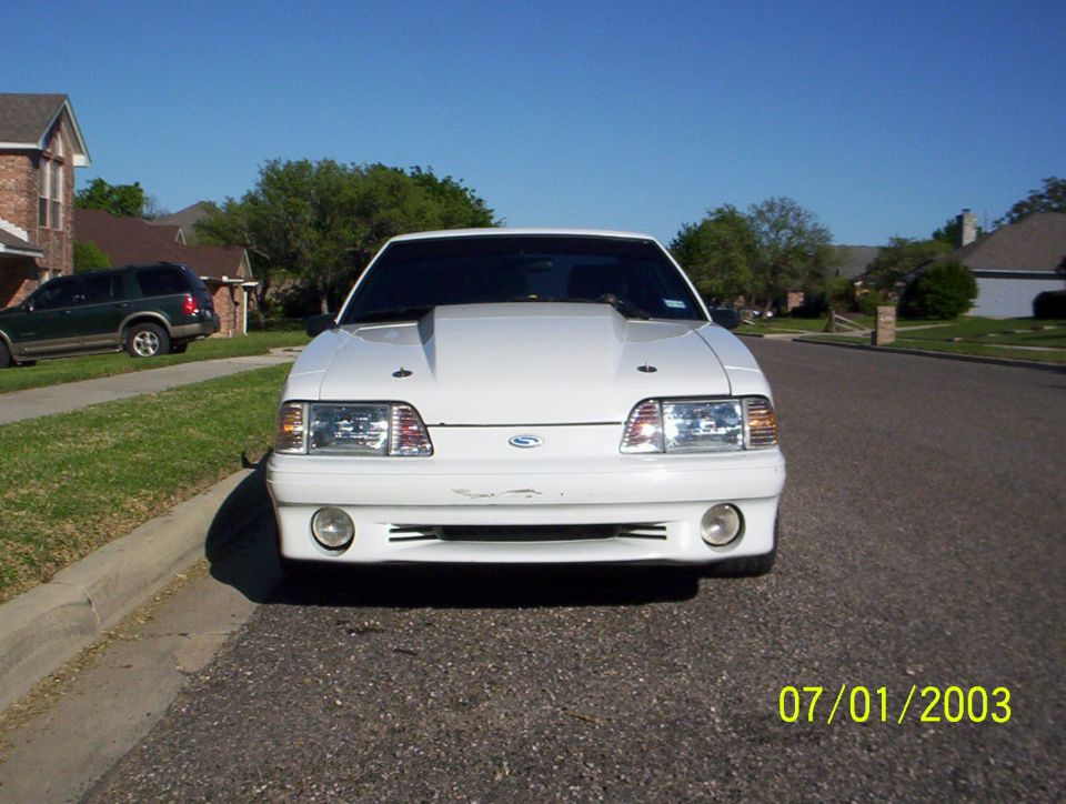 Front view of a white 1991 Ford Mustang with a raised hood, parked on a residential street