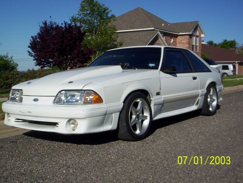 1991 Ford Mustang - A white coupe with a prominent aftermarket hood sits parked on a residential street under a clear blue sky.