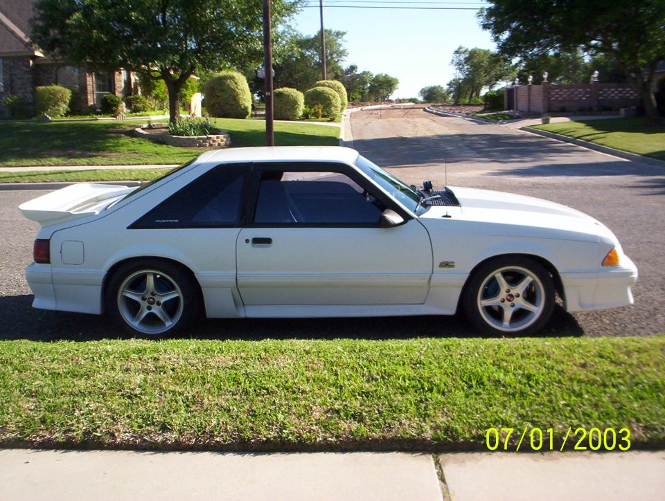 White 1991 Ford Mustang hatchback parked on a residential street, side view. Hood is slightly ajar