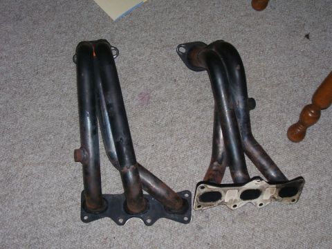 1993 Mazda MX-6 - Two exhaust manifolds are shown side-by-side on a carpeted surface. One has a black flange, the other a white gasket.