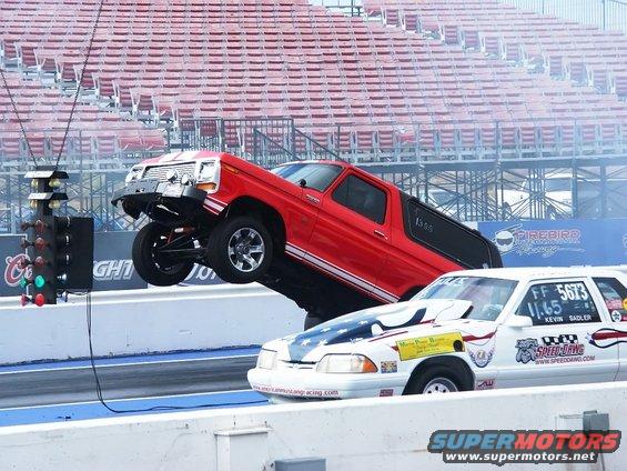 Red Ford Bronco performing a wheelie on a drag strip, with a white race car beside it