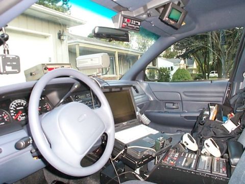 1986 Ford Crown Victoria LTD - The interior of a police vehicle, equipped with a laptop and communication devices, is shown from the driver's perspective.
