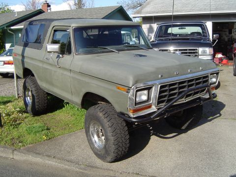 1978 Ford Bronco - A 1978 Ford Bronco with a matte olive green paint job and off-road tires sits in a driveway next to another truck.