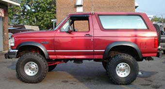 1994 Ford Bronco - Small side-profile photo of a red lifted Ford Bronco with removable hardtop, parked on asphalt in front of a brick building. It sits on ver…