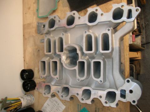 1998 Ford Crown Victoria - bare cast-aluminum lower intake manifold lies on a plywood workbench beside a green gasket and assorted tools.