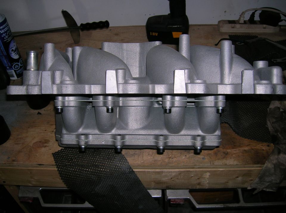 Aluminum intake manifold standing on edge on a workbench with a drill behind it