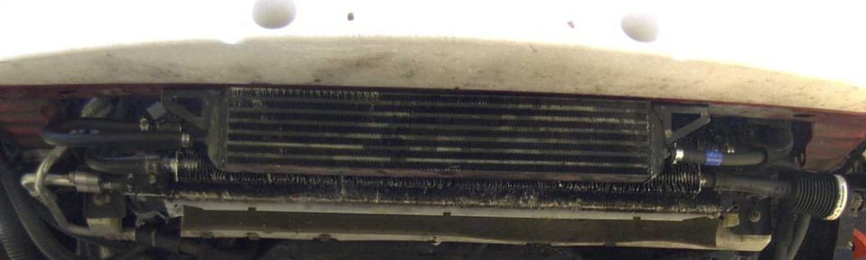 Auxiliary transmission oil cooler mounted behind a white car's front bumper with hose lines