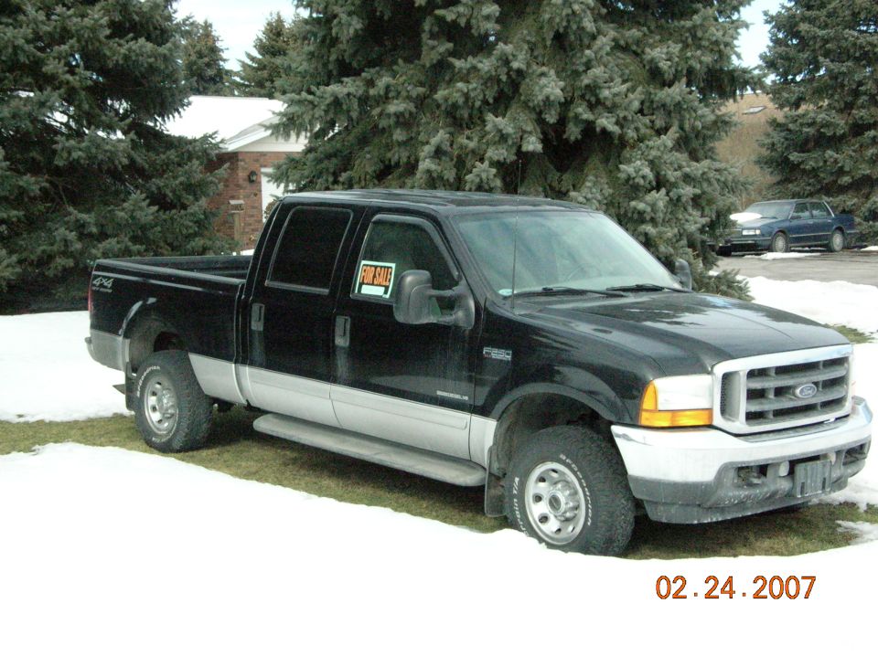 Black Ford F-250 4x4 crew cab pickup with For Sale sign, parked on snowy grass by evergreen trees