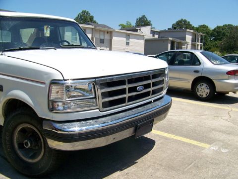 1995 Ford Bronco - A white Ford truck is parked in a lot next to a silver sedan under a clear blue sky.