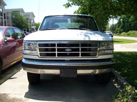 1995 Ford Bronco - A white Ford Bronco is parked in front of a building, with a red car to its left and trees overhead.