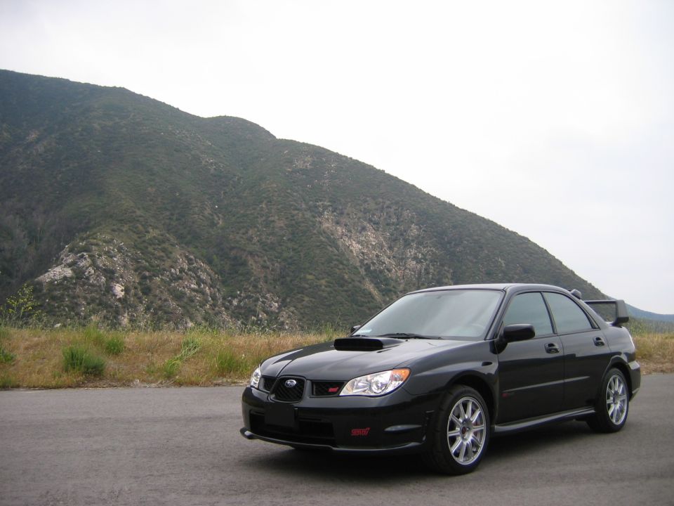 Black Subaru WRX STI sedan parked on pavement before a green mountain