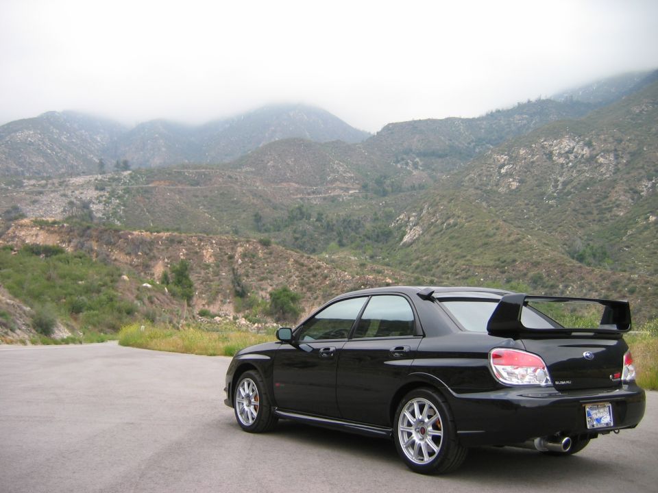 Black Subaru WRX STI sedan rear 3/4 view with large wing, parked before hazy mountains