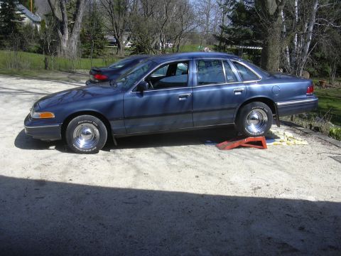 1996 Ford Crown Victoria - A blue sedan with chrome wheels is parked on ramps, with one tire appearing flat.