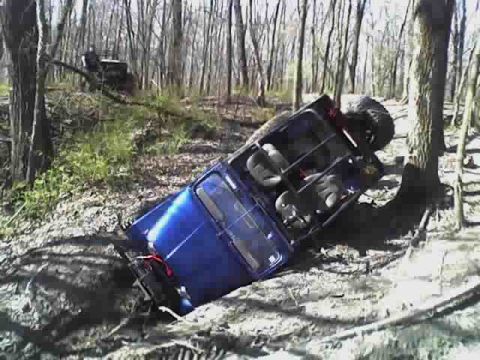 1992 Ford Bronco - An off-road vehicle has overturned on a muddy trail in a forest, with another vehicle visible in the background.