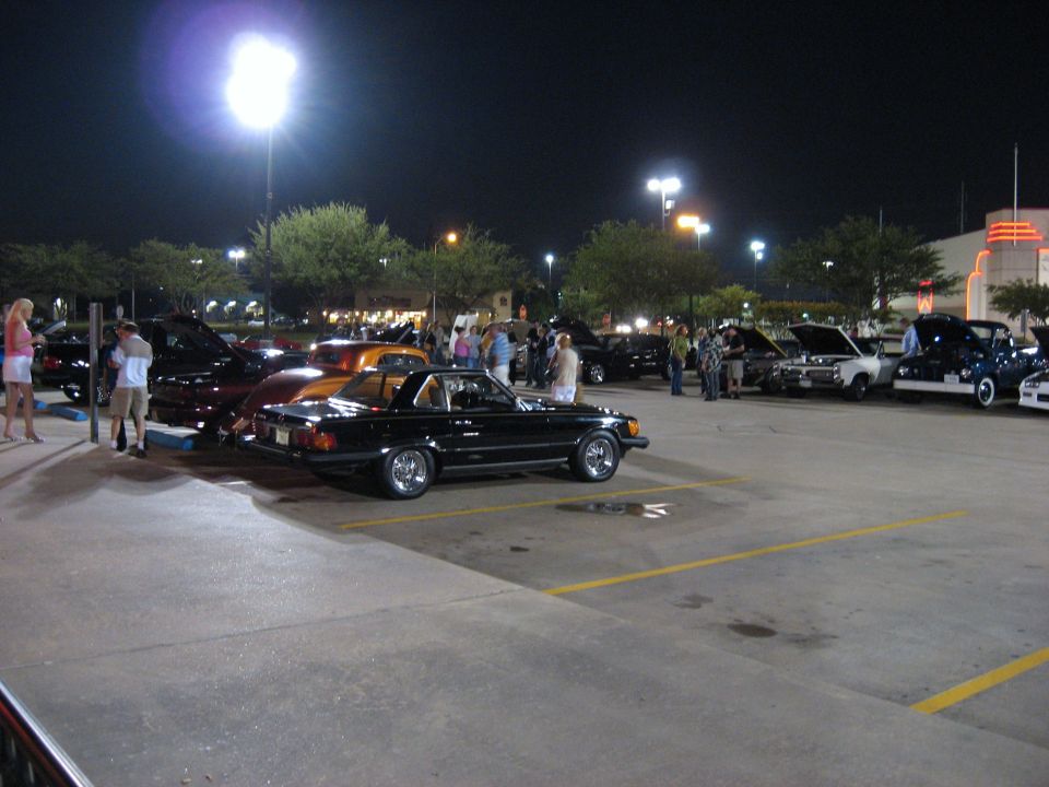 Black Mercedes SL roadster at a nighttime parking lot car show with classic cars behind