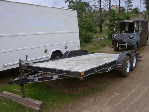 1986 Ford Bronco - An empty car trailer sits in front of a white box truck and a dark, unfinished van.