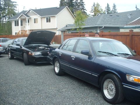 2000 Ford Crown Victoria - Two Ford Crown Victorias parked in a gravel lot, one with its hood open revealing the engine.