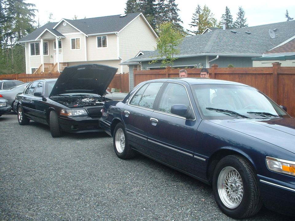Two dark blue full-size sedans on gravel, one with its hood raised as two men look on