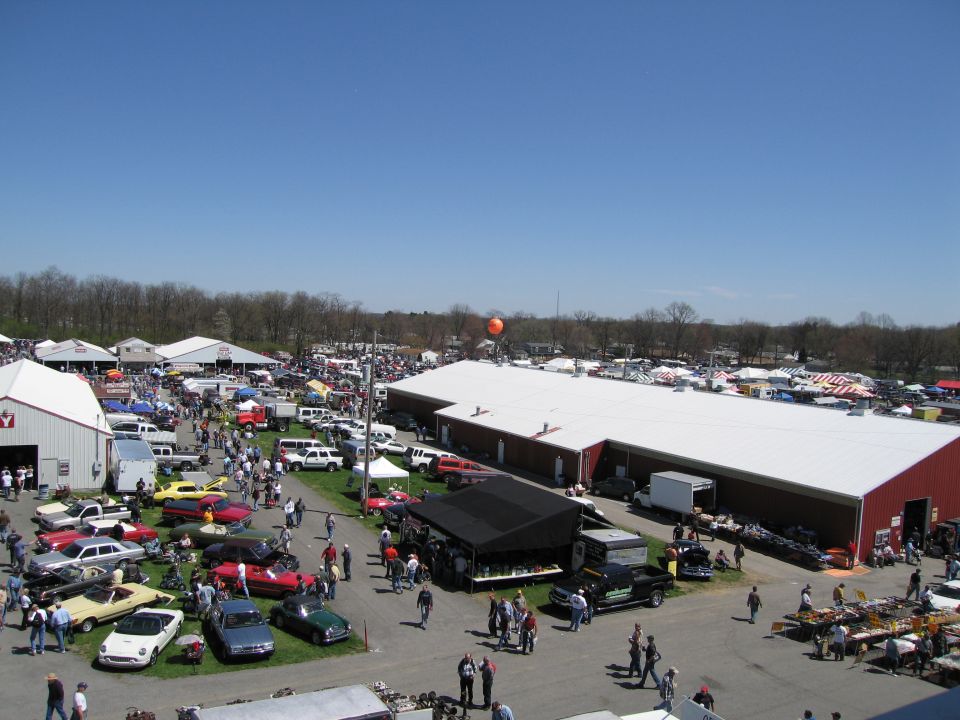 Outdoor car show and market with many vehicles and tents under a clear blue sky