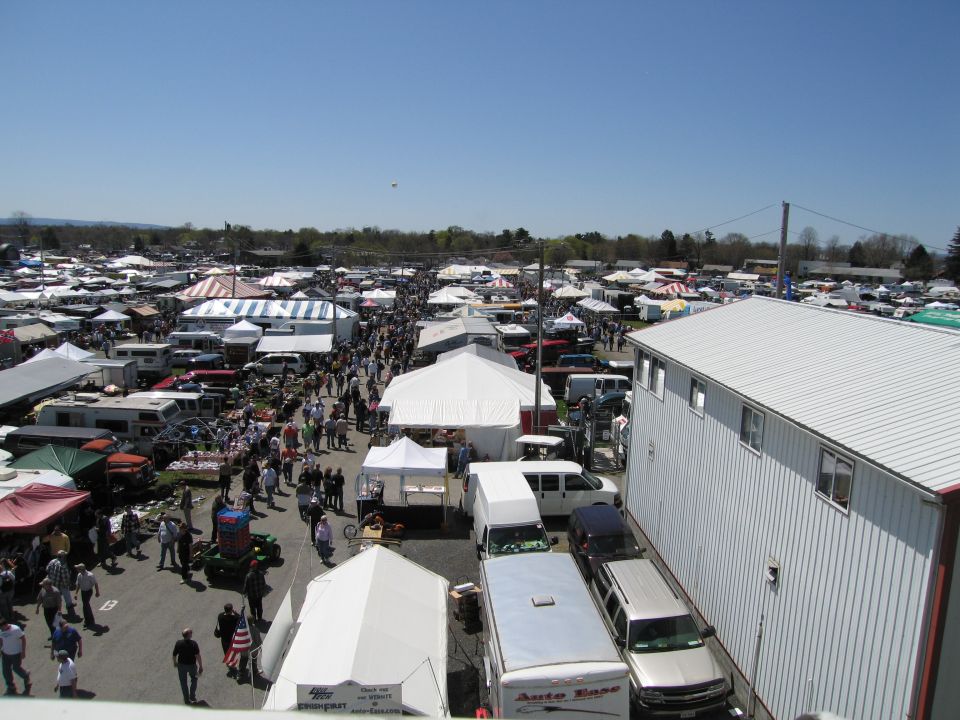 Outdoor market with rows of tents, vendors, and parked vehicles under a clear blue sky
