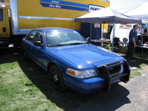 1991 Ford LTD Crown Victoria - A blue police car with a push bar is parked on the grass next to a large yellow truck.