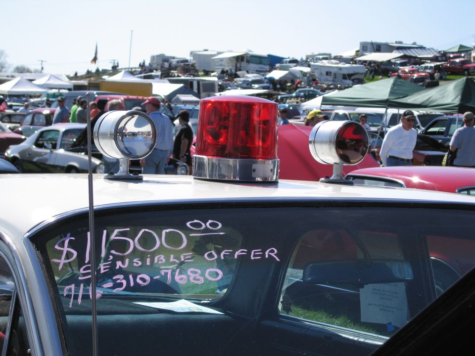 Police car roof with red light bar and spotlights, window has $1500 SENSIBLE OFFER and phone number written on it. Other cars and tents in background