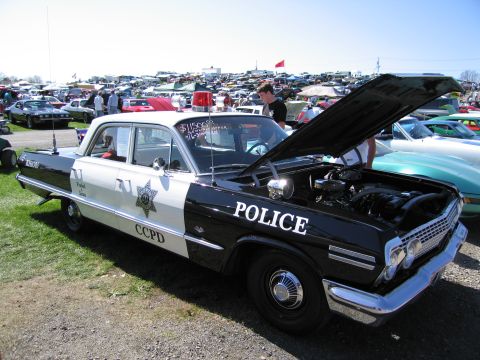 1991 Ford LTD Crown Victoria - A classic black and white police car is displayed with its hood open at a car show, surrounded by other vehicles and tents.