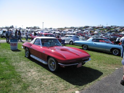 1991 Ford LTD Crown Victoria - A red 1965 Chevrolet Corvette convertible is displayed at an outdoor car show with many other vehicles in the background.