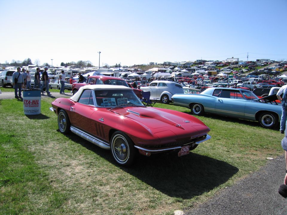 Red 1965 Chevrolet Corvette convertible with white top, parked on grass at a car show