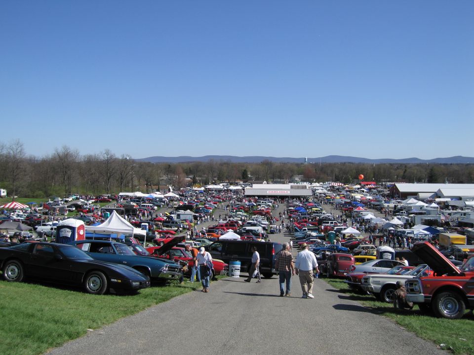 Outdoor car show with rows of cars, tents, and people under a clear blue sky