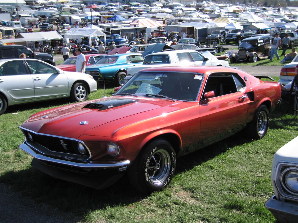 Red 1969 Ford Mustang Mach 1 parked on grass at an outdoor car show with tents and other vehicles in background