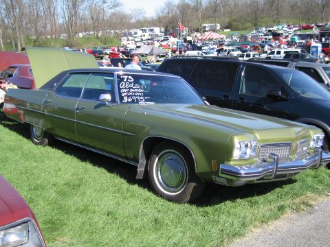 1991 Ford LTD Crown Victoria - A green 1973 Oldsmobile Delta 88 is displayed at an outdoor car show with other vehicles and tents in the background.